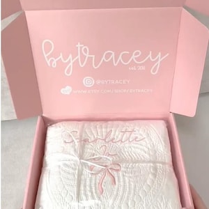 May include: A light pink cardboard shipping box with the white text bytracey est 2011, an Instagram icon with @BYTRACEY, and a website URL. Inside, a white quilted blanket is wrapped in clear plastic and tied with a white ribbon.