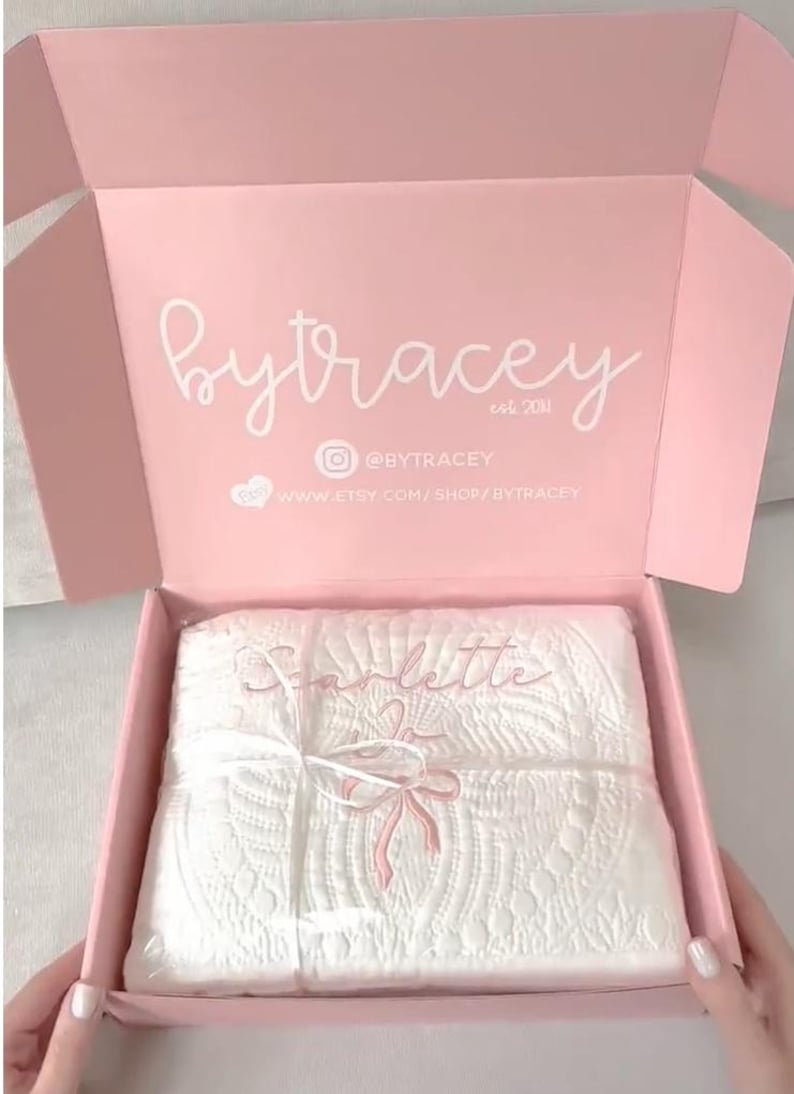 May include: A light pink cardboard shipping box with the white text bytracey est 2011, an Instagram icon with @BYTRACEY, and a website URL. Inside, a white quilted blanket is wrapped in clear plastic and tied with a white ribbon.