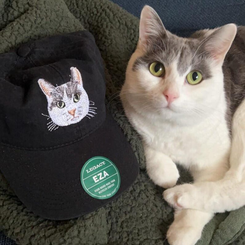 Pode incluir: Um bon&eacute; de beisebol preto com um gato branco bordado na frente. O bon&eacute; tem um adesivo verde com o texto "Legacy EZA" e as palavras "Five Star Rated Made from Recycled Content". Um gato branco e cinza est&aacute; sentado em uma superf&iacute;cie verde peluda.