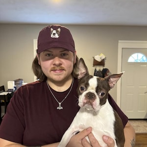 Pode incluir: Uma pessoa usando um bon&eacute; de beisebol vinho com um cachorro branco e preto bordado na frente. A pessoa est&aacute; segurando um Boston Terrier branco e marrom.