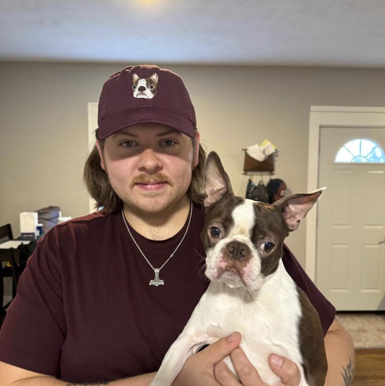 Pode incluir: Uma pessoa usando um bon&eacute; de beisebol vinho com um cachorro branco e preto bordado na frente. A pessoa est&aacute; segurando um Boston Terrier branco e marrom.