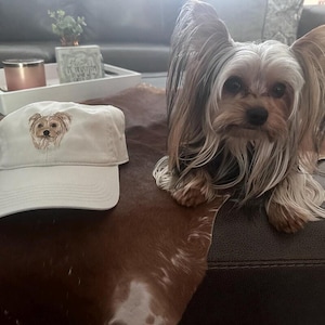 Op de afbeelding: Een witte baseballpet met een geborduurd beeld van een Yorkshire Terrier. De hond is bruin en beige met zwarte ogen en een zwarte neus. De pet ligt op een bruin en wit koeienhuidkleed.