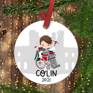 Puede incluir: Adorno de Navidad redondo blanco con una ilustración de dibujos animados de un niño en silla de ruedas con un traje de caballero y sosteniendo una espada. El niño está frente a una silueta de castillo gris. El adorno tiene el nombre "COLIN" y el año "2021" impreso en él.