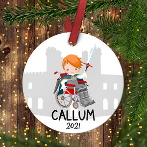 Puede incluir: Adorno navideño redondo blanco con una ilustración de dibujos animados de un niño en silla de ruedas con un traje de caballero y sosteniendo una espada. El adorno tiene una silueta de castillo gris en el fondo y el texto "CALLUM 2021".