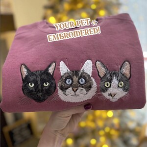 Op de afbeelding: Een mauvekleurige sweatshirt met drie geborduurde kattengezichten. De katten zijn zwart, bruin en wit, en grijs en wit. De tekst "YOUR PET EMBROIDERED!" staat boven de kattengezichten.