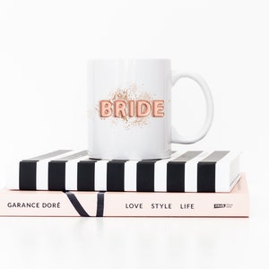 Puede incluir: Taza de cer&aacute;mica blanca con un gr&aacute;fico "BRIDE" en oro rosa sobre fondo blanco. La taza est&aacute; encima de dos libros, uno con rayas blancas y negras y el otro con el texto "GARANCE DOR&Eacute;" y "LOVE STYLE LIFE".