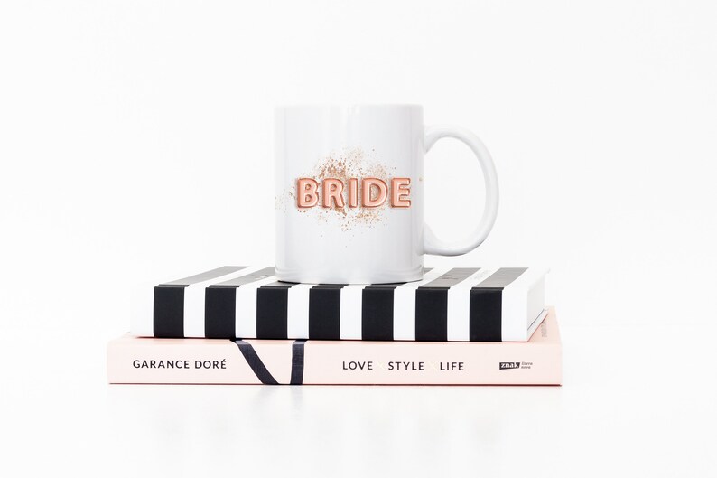 Puede incluir: Taza de cer&aacute;mica blanca con un gr&aacute;fico "BRIDE" en oro rosa sobre fondo blanco. La taza est&aacute; encima de dos libros, uno con rayas blancas y negras y el otro con el texto "GARANCE DOR&Eacute;" y "LOVE STYLE LIFE".
