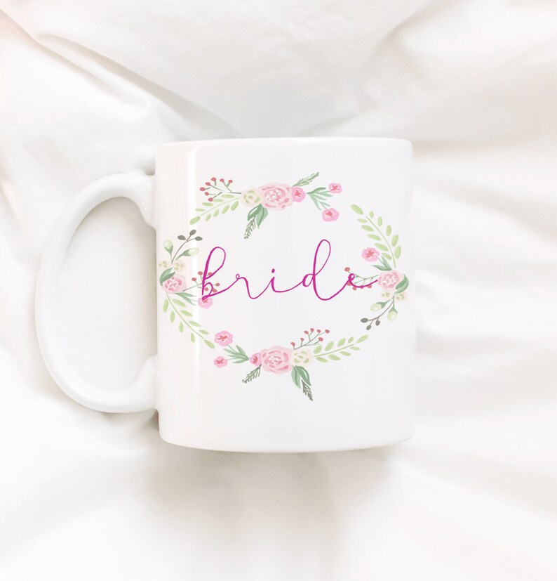 Pode incluir: Caneca de cer&acirc;mica branca com um design de coroa de flores e a palavra "bride" em script rosa. A coroa apresenta rosas cor-de-rosa, folhas verdes e bagas vermelhas. A caneca tem uma al&ccedil;a grande e curva.