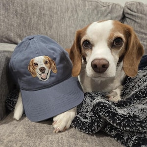 Op de afbeelding: Een blauwe baseballpet met een geborduurd beeld van een beagle. De hond kijkt recht in de camera met zijn tong naar buiten. De pet ligt op een bank naast een beagle.