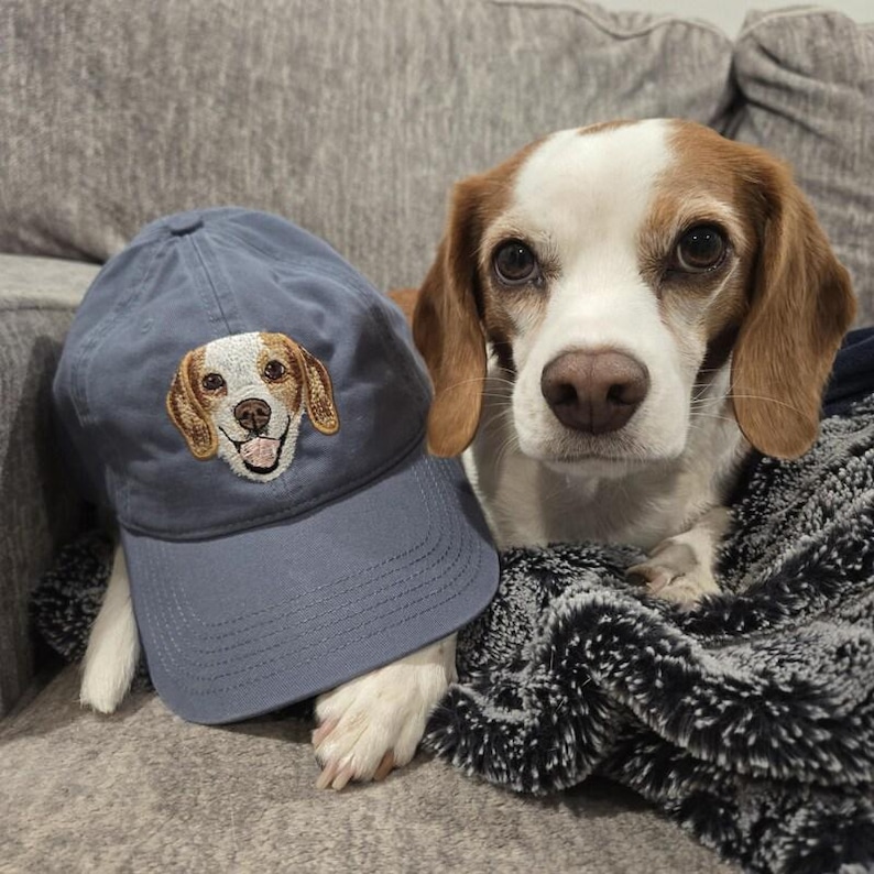 Op de afbeelding: Een blauwe baseballpet met een geborduurd beeld van een beagle. De hond kijkt recht in de camera met zijn tong naar buiten. De pet ligt op een bank naast een beagle.