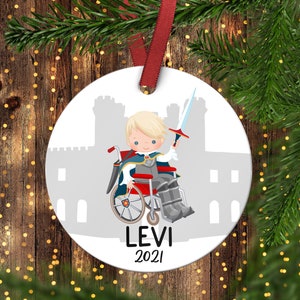 Puede incluir: Adorno navideño blanco redondo con una ilustración de dibujos animados de un joven caballero en silla de ruedas, sosteniendo una espada. El adorno tiene una cinta roja para colgar y el nombre "LEVI" y el año "2021". Una silueta de castillo gris en el fondo.