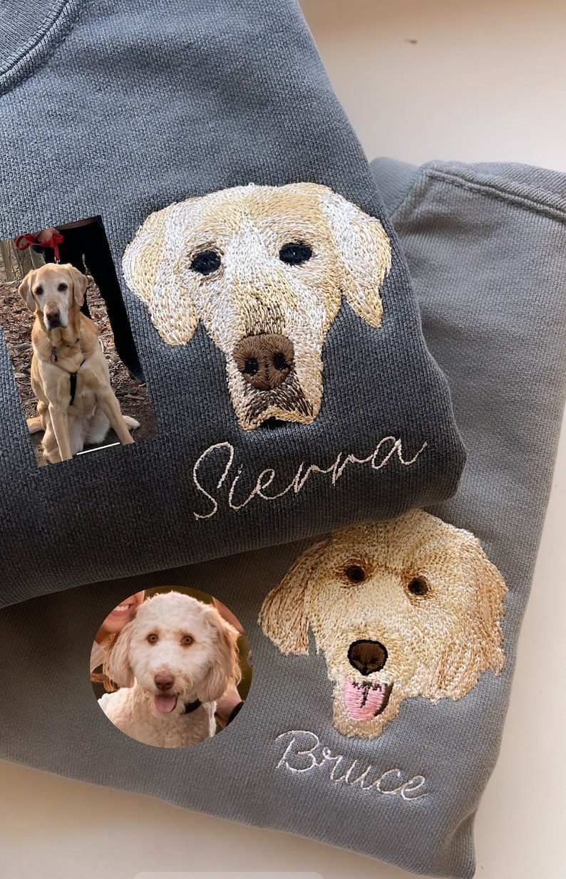 Op de afbeelding: Twee grijze sweatshirts met op maat geborduurde hondenportretten. E&eacute;n sweatshirt heeft een portret van een golden retriever met de naam "Sierra" eronder geborduurd. De andere heeft een vergelijkbaar portret met de naam "Bruce".