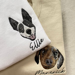 Op de afbeelding: Twee sweatshirts, &eacute;&eacute;n witte en &eacute;&eacute;n beige, elk geborduurd met het gezicht en de naam van een hond. Het witte sweatshirt heeft een zwart-witte hond genaamd "Ellie". Het beige sweatshirt heeft een bruine hond genaamd "Maverick".