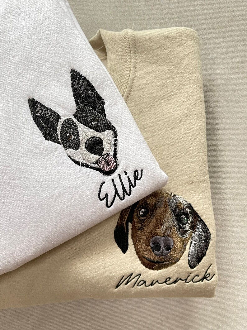 Op de afbeelding: Twee sweatshirts, &eacute;&eacute;n witte en &eacute;&eacute;n beige, elk geborduurd met het gezicht en de naam van een hond. Het witte sweatshirt heeft een zwart-witte hond genaamd "Ellie". Het beige sweatshirt heeft een bruine hond genaamd "Maverick".