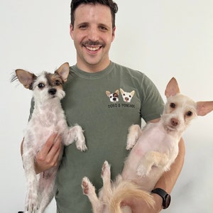 Op de afbeelding: Een man draagt een groen t-shirt met de tekst "TOTO & PONCHOS" en twee honden gezichten. Hij houdt twee kleine honden vast, een met bruin en wit haar, de andere met wit haar. De achtergrond is effen wit.
