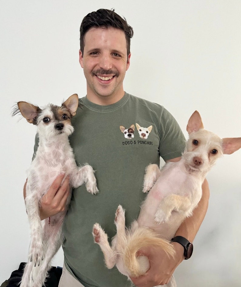 Op de afbeelding: Een man draagt een groen t-shirt met de tekst "TOTO & PONCHOS" en twee honden gezichten. Hij houdt twee kleine honden vast, een met bruin en wit haar, de andere met wit haar. De achtergrond is effen wit.