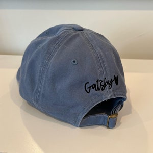 Op de afbeelding: Een blauwe denim baseballpet met zwarte borduurwerk die "Gatsby" met een hart zegt.