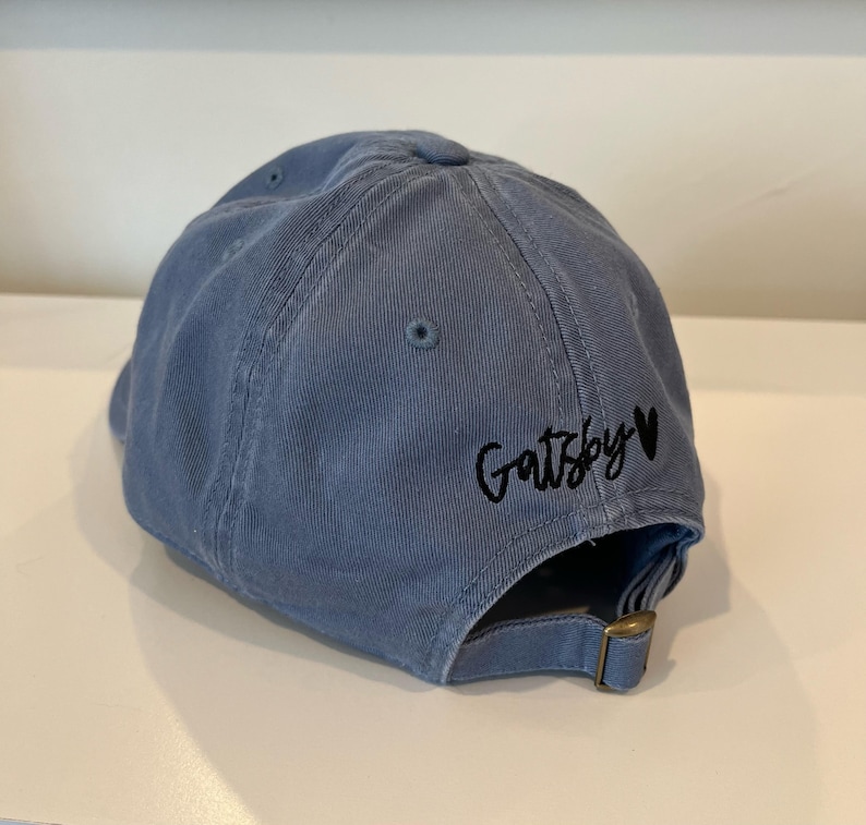 Op de afbeelding: Een blauwe denim baseballpet met zwarte borduurwerk die "Gatsby" met een hart zegt.