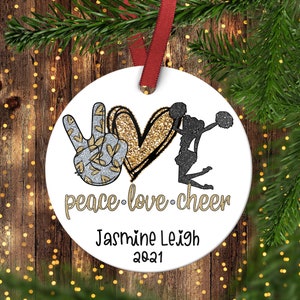 May include: A white round Christmas ornament with a peace sign, a gold glitter heart, and a black silhouette of a cheerleader. The text "peace love cheer" is written below the heart. The ornament is personalized with the name "Jasmine Leigh" and the year "2021".