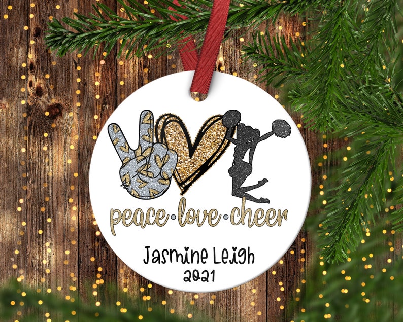 May include: A white round Christmas ornament with a peace sign, a gold glitter heart, and a black silhouette of a cheerleader. The text "peace love cheer" is written below the heart. The ornament is personalized with the name "Jasmine Leigh" and the year "2021".