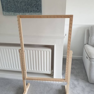 Op de afbeelding: Een houten weefgetouw met een rechthoekig frame en metalen pinnen. Het weefgetouw staat op een tapijt in de voorkant van een witte radiator.