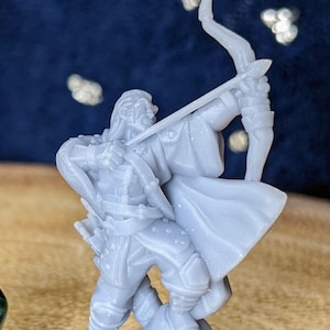 May include: A gray miniature figurine of an archer with a bow and arrow, standing on a rocky base. A green and gold twenty-sided die is next to the figurine. The background is a dark blue with bokeh lights.