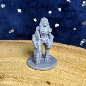 May include: A grey 3D printed miniature of a futuristic soldier wearing a hooded cloak and holding a weapon. The miniature is standing on a round base.