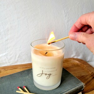 May include: A clear glass candle jar with a white label that reads "In thy presence is fullness of Joy, Psalm 16:11". The candle is lit with a match.