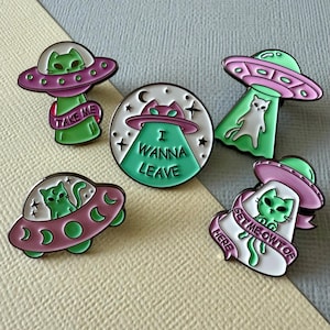May include: Five enamel pins with cat-themed alien spaceship designs. The pins are in green, pink, and white. Text includes "Take Me" and "I Wanna Leave". A quirky accessory for bags or clothing. The pins are a fun and quirky accessory.