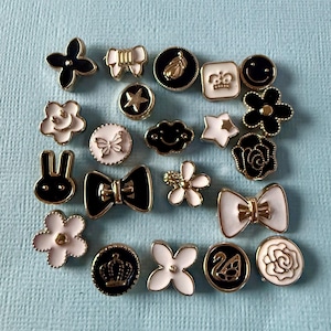 May include: A selection of decorative buttons in diverse shapes, such as flowers, bows, stars, and a bunny. The buttons showcase black and white enamel designs with gold accents, set against a light blue backdrop.