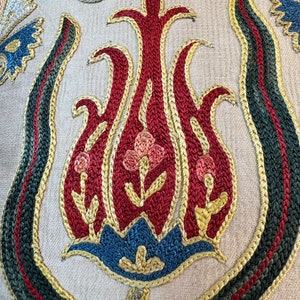 May include: Close-up of a detailed embroidered design featuring a stylized tulip with red, pink, blue, and gold thread on a beige fabric background.