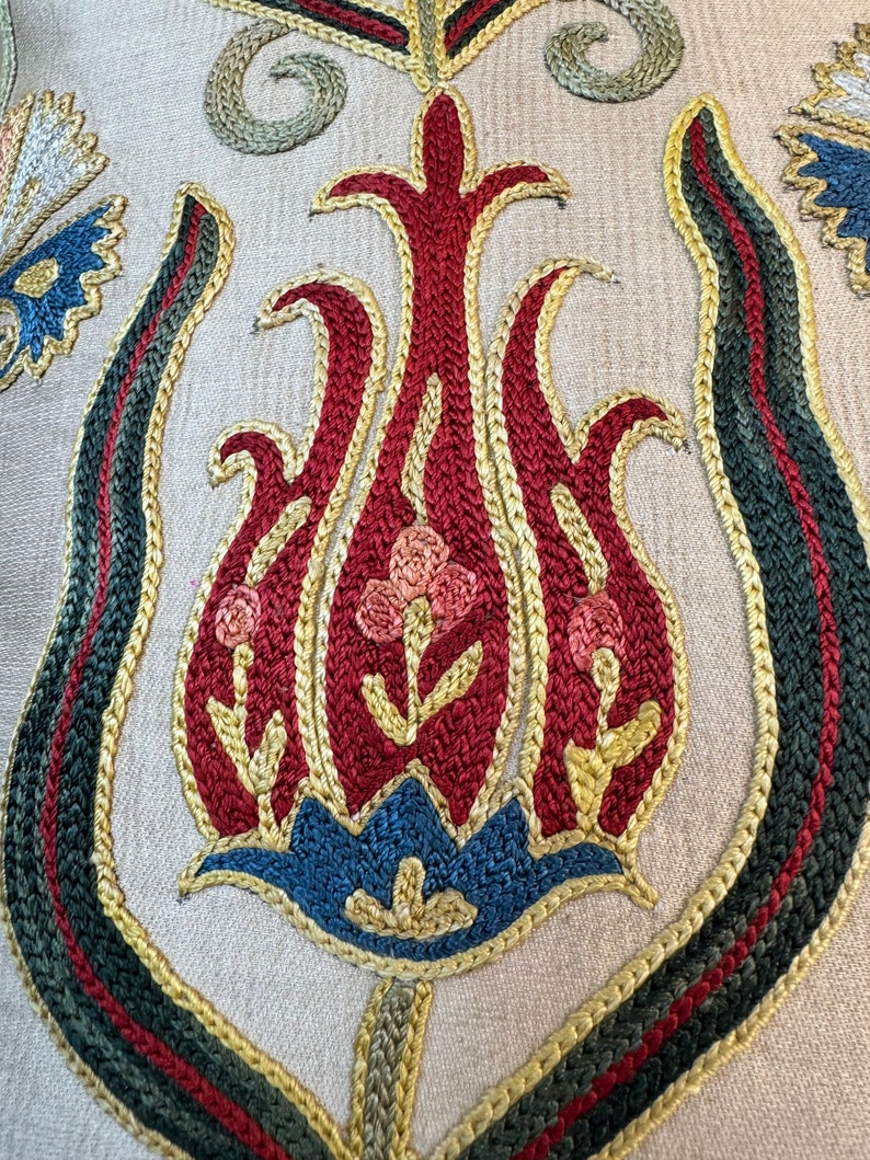 May include: Close-up of a detailed embroidered design featuring a stylized tulip with red, pink, blue, and gold thread on a beige fabric background.
