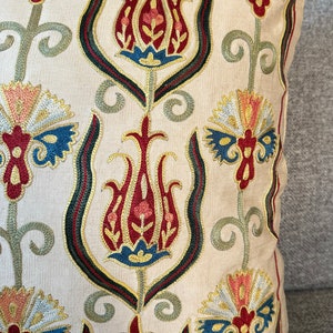 May include: A cream-colored pillow with a traditional Turkish design embroidered in red, blue, green, and gold thread. The design features stylized flowers and leaves.