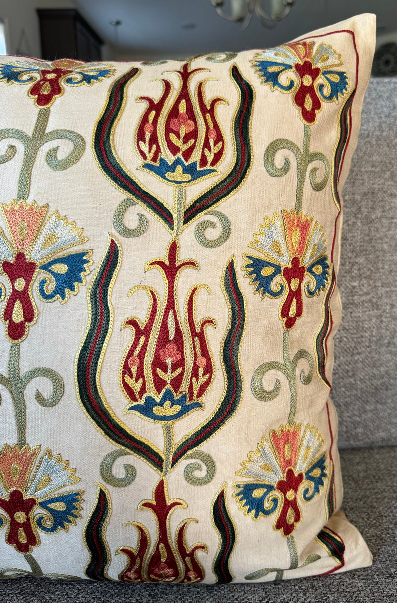 May include: A cream-colored pillow with a traditional Turkish design embroidered in red, blue, green, and gold thread. The design features stylized flowers and leaves.