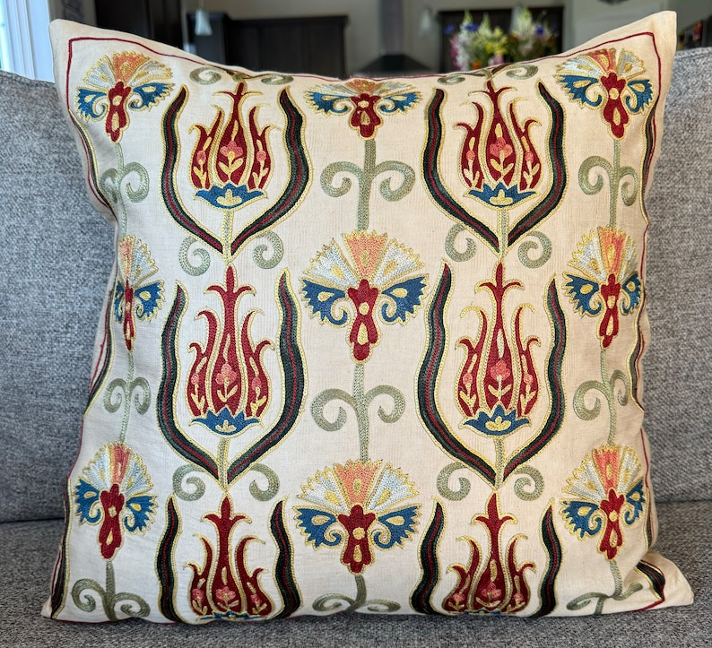 May include: A cream-colored throw pillow with a repeating pattern of embroidered red, blue, and green stylized tulips. The pillow has a subtle sheen and a soft, textured fabric.