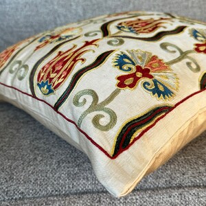 May include: A cream-colored pillow with embroidered floral designs in shades of red, green, blue, and gold. The pillow has a decorative border with a dark green, black, and red trim.
