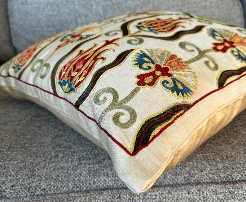 May include: A cream-colored pillow with embroidered floral designs in shades of red, green, blue, and gold. The pillow has a decorative border with a dark green, black, and red trim.