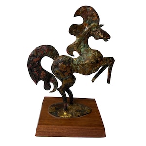 May include: A copper-toned metal sculpture of a rearing horse, mounted on a rectangular wooden base. The horse has a stylized mane and tail, with a textured, aged appearance. The sculpture is a decorative art piece.