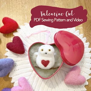 Puede incluir: Un búho de fieltro blanco con un corazón rojo en el pecho se sienta dentro de una lata roja en forma de corazón. La lata está abierta y el búho está mirando hacia afuera. La lata está sobre un encaje de ganchillo blanco. Hay varios corazones de fieltro rojo, rosa y morado alrededor de la lata. El texto "Valentine Owl PDF Sewing Pattern and Video" está escrito en letras blancas sobre un fondo rojo.