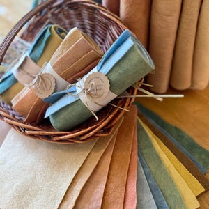 Op de afbeelding: Een verzameling gekleurde vellen en rollen vilt in verschillende aardetinten. Gerolde viltbundels zijn vastgebonden met touw en papieren labels, gepresenteerd in een rieten mand. Losse vellen waaieren eronder uit en tonen de reeks kleuren.