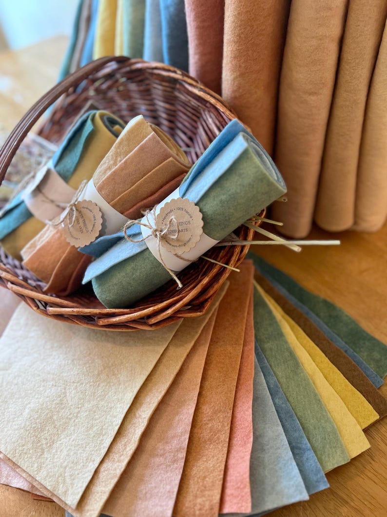 Op de afbeelding: Een verzameling gekleurde vellen en rollen vilt in verschillende aardetinten. Gerolde viltbundels zijn vastgebonden met touw en papieren labels, gepresenteerd in een rieten mand. Losse vellen waaieren eronder uit en tonen de reeks kleuren.