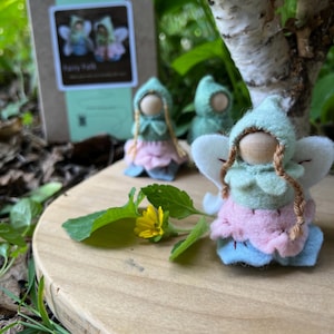 May include: Three small peg dolls with wooden heads, wearing felt hoods, light green tops, pink skirts, and blue bases, featuring white felt wings and braided yarn hair. A small cardboard box with a label reading Fairy Folk is visible in the background.