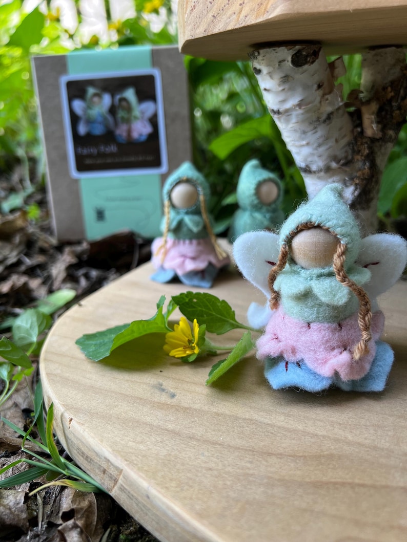 May include: Three small peg dolls with wooden heads, wearing felt hoods, light green tops, pink skirts, and blue bases, featuring white felt wings and braided yarn hair. A small cardboard box with a label reading Fairy Folk is visible in the background.