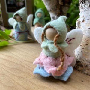 May include: A small felt fairy figurine with a light green pointed hood, a pink petal-shaped skirt, and light blue base layers. The figure has a round wooden head, two thin brown braided strands, and white felt wings with red stitching details.