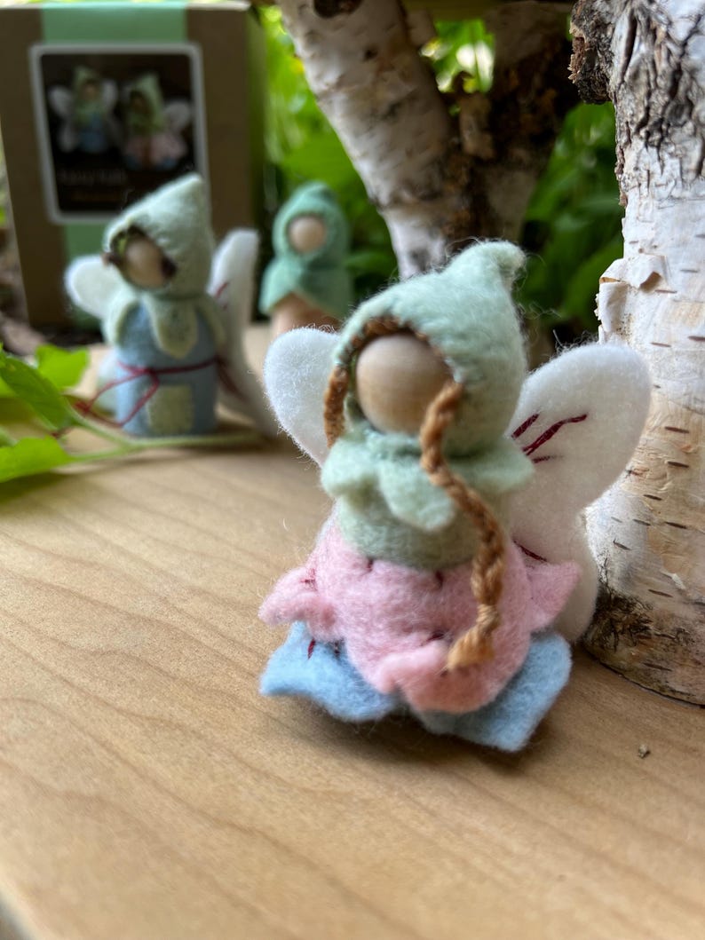 May include: A small felt fairy figurine with a light green pointed hood, a pink petal-shaped skirt, and light blue base layers. The figure has a round wooden head, two thin brown braided strands, and white felt wings with red stitching details.