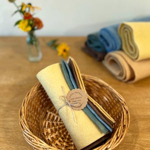 Op de afbeelding: Een rieten mand bevat een bundel gele viltvellen vastgebonden met touw, naast verschillende andere opgerolde viltvellen in tinten blauw, bruin en beige. Een kleine vaas met bloemen staat op de achtergrond. Het label luidt "Wee Things Fiber Arts."