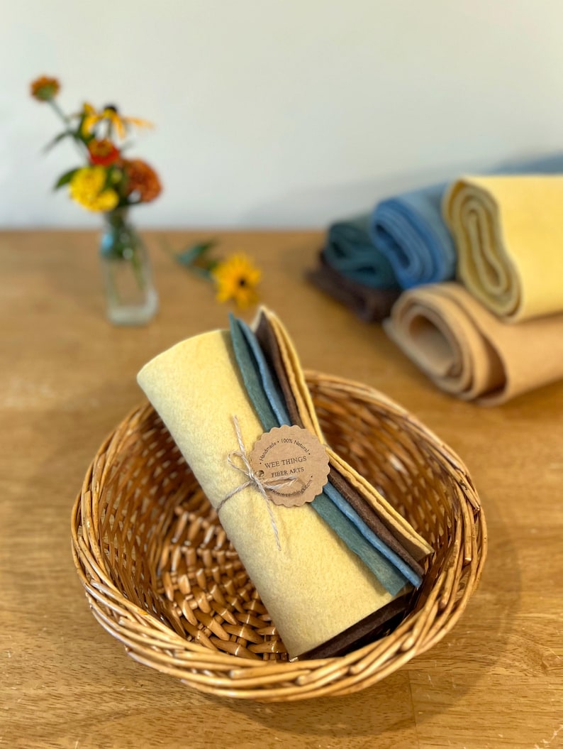 Op de afbeelding: Een rieten mand bevat een bundel gele viltvellen vastgebonden met touw, naast verschillende andere opgerolde viltvellen in tinten blauw, bruin en beige. Een kleine vaas met bloemen staat op de achtergrond. Het label luidt "Wee Things Fiber Arts."