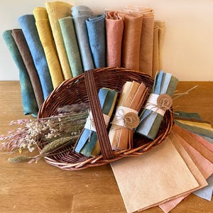 Op de afbeelding: Een rieten mand volgepakt met gekleurde stoffen rollen en bundels. De rollen zijn in tinten blauw, geel, bruin en perzik. Bundels zijn vastgebonden met touw en papieren labels. Een selectie platte stoffen vierkanten in vergelijkbare kleuren is uitgespreid.