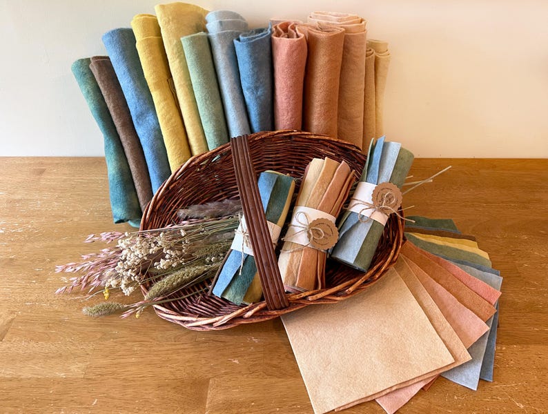 Op de afbeelding: Een rieten mand volgepakt met gekleurde stoffen rollen en bundels. De rollen zijn in tinten blauw, geel, bruin en perzik. Bundels zijn vastgebonden met touw en papieren labels. Een selectie platte stoffen vierkanten in vergelijkbare kleuren is uitgespreid.