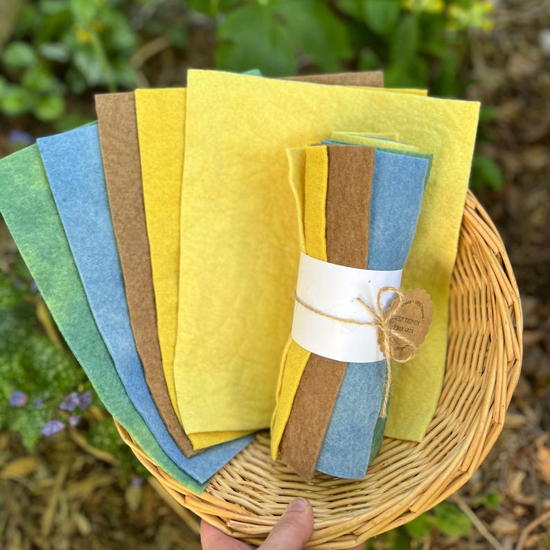 Op de afbeelding: Een verzameling kleurrijke vellen vilt in groen, blauw, bruin en geel, gerangschikt in een rieten mand. Sommige vellen zijn gebundeld met een witte band en touw, met een klein label eraan. Knutselbenodigdheden.